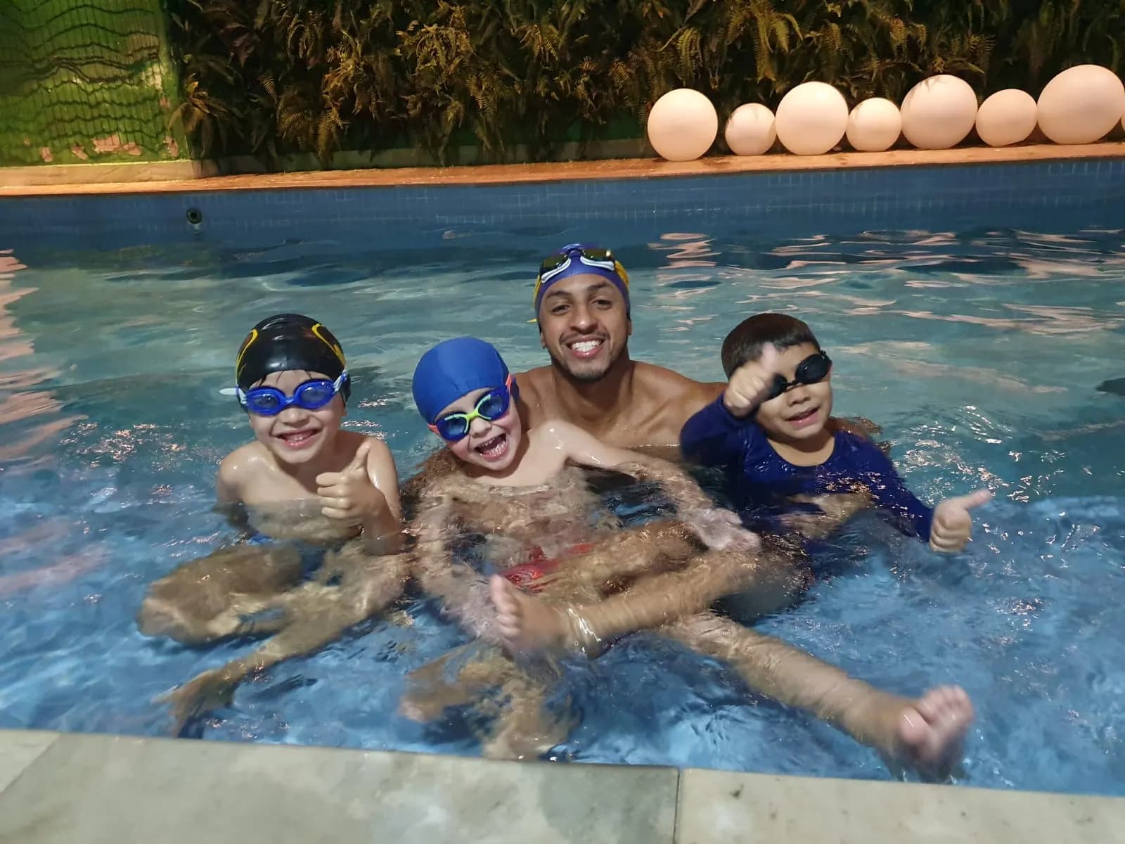 Professor dentro da piscina com três meninos de touca e óculos fazendo sinal de positivo